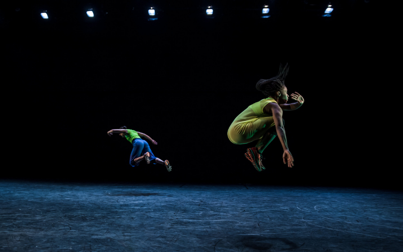 Two dancers in neon clothing jump on a dark stage