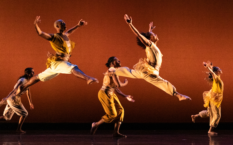 Five dancers in yellow costumes leaping in the air against a red orange background