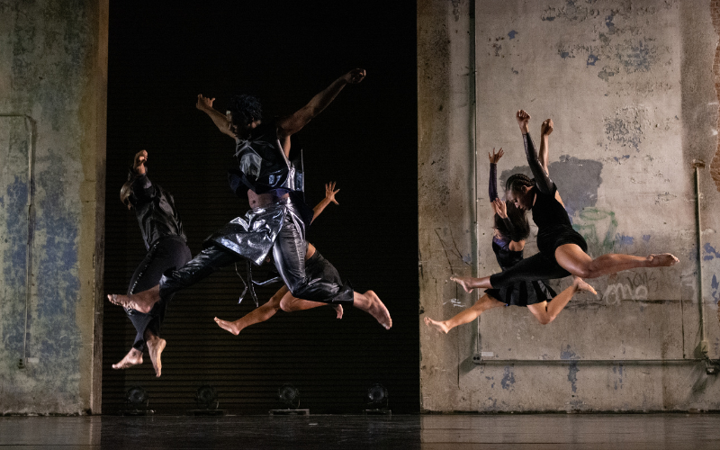 5 dancers leap through the air in an industrial setting.