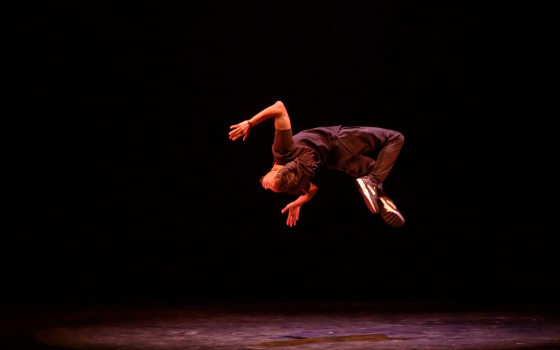 A dancer suspended mid-air during a back flip on a dark stage.