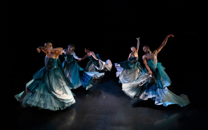 5 dancers wearing flowing blue dresses form a V on stage.