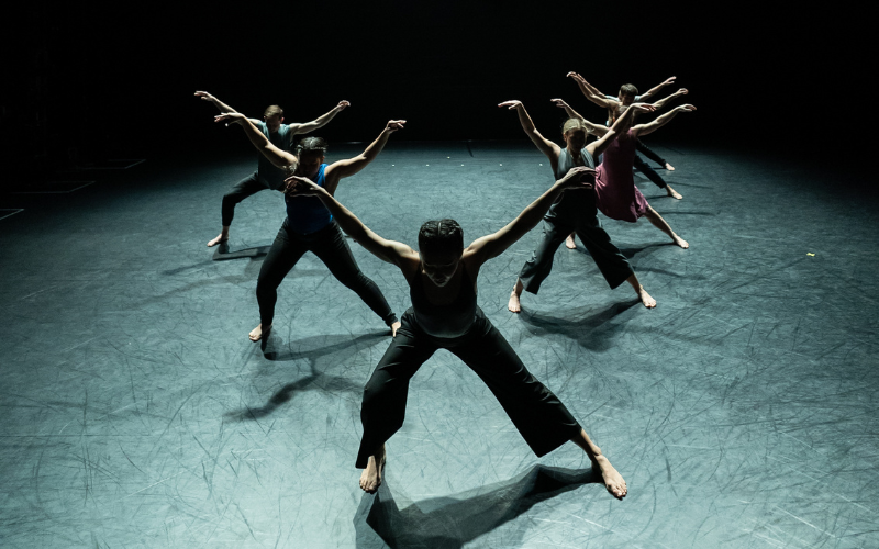 6 dancers form a V on stage with their legs in a lunge and their arms outstretched wide.